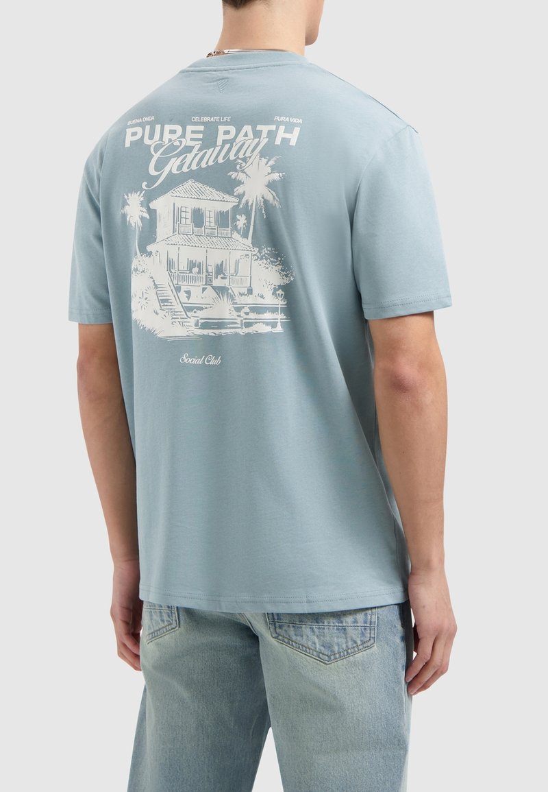 Lichtblauw katoenen t-shirt met een witte afbeelding van een huis en palmbomen op de achterkant. Relaxed fit met korte mouwen en een ronde hals.