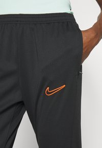 Pantalones deportivos negros con cintura elástica, que presentan un logotipo de Nike bordado en naranja en el muslo izquierdo. Tejido liso y ligero.