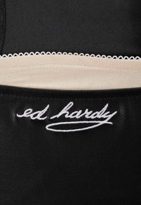 Material sintético negro con un acabado texturizado, que presenta un logo bordado en blanco de "ed hardy" y un acento crema con un borde en forma de doble curva.
