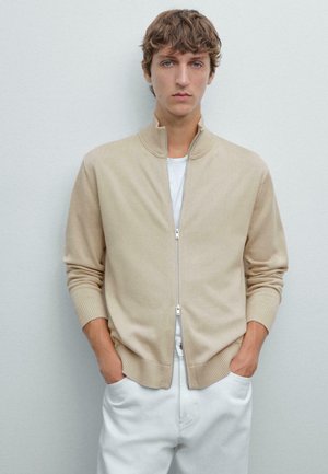 Cardigan beige con zip, colletto alto, polsini e orlo a coste, in un tessuto a maglia leggero, indossato sopra una t-shirt bianca e abbinato a pantaloni bianchi.