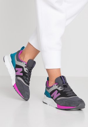 Person wearing gray, black, teal, and purple New Balance sneakers with white soles, paired with white pants against a plain background.