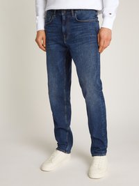 Jeans de ganga azul escuro com cinco bolsos, fecho de correr, costura laranja e corte afunilado, combinados com ténis brancos.