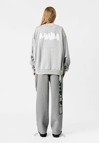 Sudadera gris y pantalones de chándal a juego, con un logo en la parte trasera y detalles en los Laterales. Corte holgado, con puños acanalados y dobladillos ajustables con cordón.