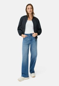 Frau in einem weißen Tanktop, schwarzer Bomberjacke, weiten blauen Jeans und weißen Sneakers, die mit Händen in den Taschen dasteht.