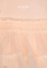 Brzoskwiniowa sukienka z miękkim bawełnianym gorsetem i warstwową spódnicą z tiulu, ozdobiona marszczeniami oraz logo marki "GUESS EST. 1981" w białym kolorze.