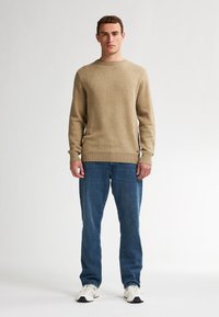 Beige gebreide trui met een ronde hals, lange mouwen en geribde manchetten, gecombineerd met blauwe jeans en witte sneakers, staand tegen een effen achtergrond.