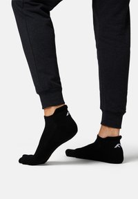 Black ankle socks with a textured design and a white logo on the heel, paired with dark gray joggers, displaying a close-up view of the feet.