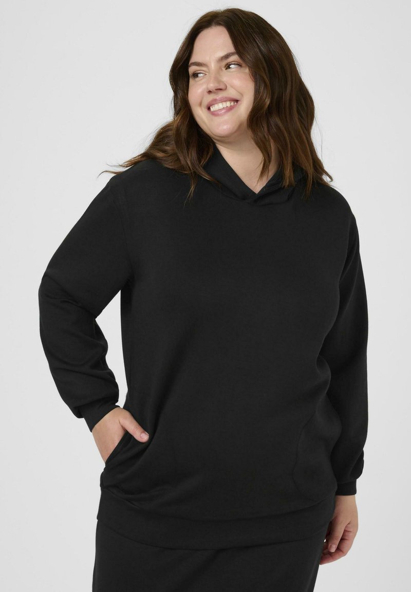 Femme souriante portant un sweat à capuche noir à manches longues, avec une main dans sa poche, regardant sur le côté devant un fond neutre.