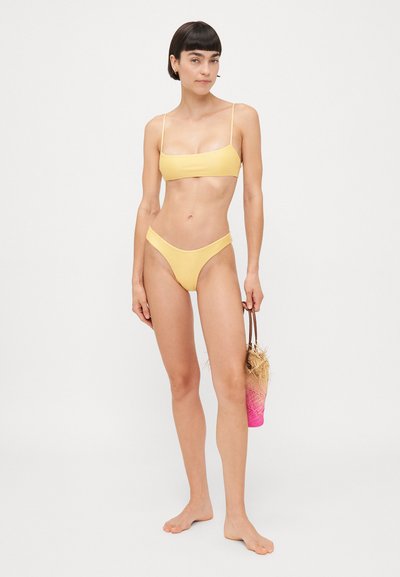 Yellow bikini set with thin straps. The top is a bandeau style, and the bottom features a high-cut design. Accompanied by a pink and straw bag.