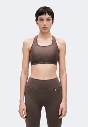Donna che indossa un reggiseno sportivo marrone Under Armour e leggings, in piedi di fronte a uno sfondo chiaro e uniforme.