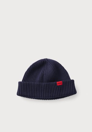 Navy knit beanie with a foldable ribbed cuff. Features a small red brand patch on the side. Made of soft, textured material.