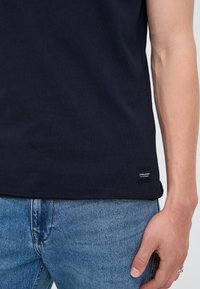 A person wearing a black t-shirt with a small logo tag near the bottom hem and blue denim jeans, with one arm relaxed at their side.