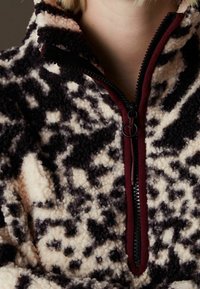 Kurtka polarowa z czarno-białym abstrakcyjnym wzorem i bordowym zamkiem błyskawicznym na górnej części tułowia i szyi osoby.