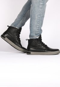 Černé kožené vysoké tenisky s šedou gumovou podrážkou, kulatou špičkou, šesti dírkami na šňůrky, texturovaným povrchem a poutkem na patě pro snadné obouvání.