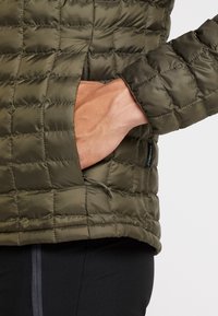 Olivgrön puffjacka med ett quiltat mönster, som har en dragkedjeficka och elastiska ärmslut. Slät textur och lättviktsdesign.