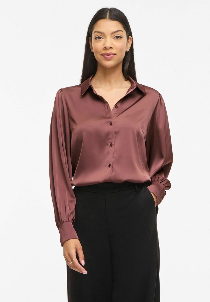 Mujer con blusa de manga larga de satén en color malva oscuro con botones y pantalones negros, de pie con una mano en el bolsillo contra un fondo blanco.