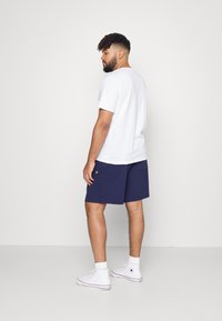 Hvid T-shirt med korte ærmer, marineblå shorts med gult logo, hvide høj-top sneakers, stående model set fra ryggen mod en lys baggrund.