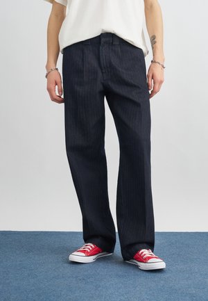 Személy sötét, széles szárú farmernadrágot, fehér rövid ujjú inget, piros alacsony szárú sportcipőt és ezüst karkötőket visel mindkét csuklóján.