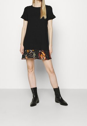 Woman wearing a black short-sleeve dress with a colorful floral hem, paired with black ankle boots, standing with legs slightly apart.