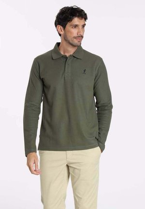 Polo shirt à manches longues vert foncé avec un col classique et un plastron à trois boutons. Avec logo brodé sur la poitrine. Associé à un pantalon beige.