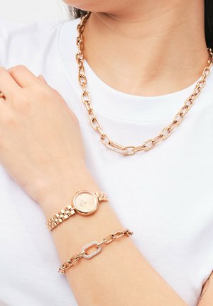 Guldfärgad smycken på en person, inklusive ett kraftigt kedjehalsband, en klocka med rund urtavla och ett armband med en länk med accenter.
