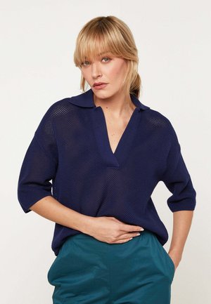 Frau mit blondem Haar, die einen marineblauen Stricktop mit offenem Kragen und türkisfarbene Hose trägt, posiert mit einer Hand in der Tasche vor weißem Hintergrund.