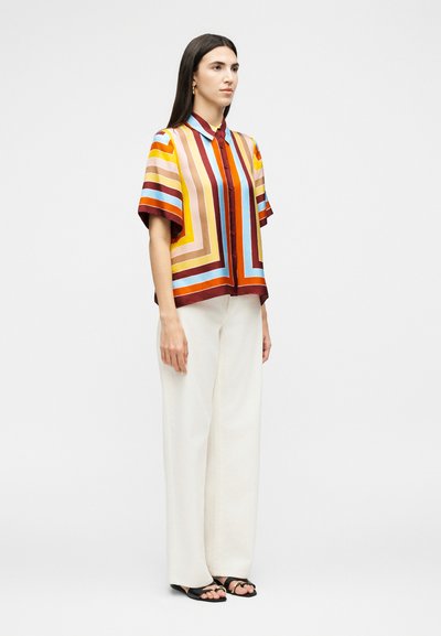 Γυναίκα που στέκεται, φοράει πολύχρωμο ριγέ πουκάμισο με κοντά μανίκια και κουμπιά, λευκό παντελόνι με φαρδιά πόδια και μαύρα σανδάλια με ανοιχτά δάχτυλα σε απλό φόντο.