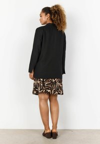 Schwarzer, leichter Blazer mit langen Ärmeln, kombiniert mit einem braunen, gemusterten Rock mit Blättermotiven. Zu sehen sind braune, hochhackige Schuhe und lockiges Haar.