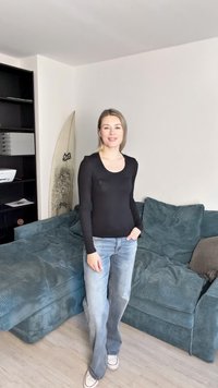 Fekete, hosszú ujjú póló, kék farmernadrág, egy textúrázott, türkiz kék kanapén áll, előtérben egy szörfdeszkával, világos fa padló.