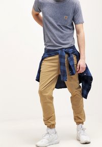 Mężczyzna w khaki joggerach, niebieskiej koszuli w kratę związanej w talii, szarym t-shircie i białych trampkach stoi na tle białym.