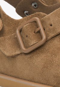 Brown suede shoe with a flat sole, featuring a rectangular brown buckle and stitching details. The material has a soft, textured surface.