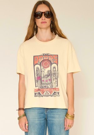 Femme portant un t-shirt graphique crème avec un motif vintage et un jean bleu, accessoirisée de grandes lunettes de soleil, de boucles d'oreilles et de multiples bracelets.