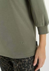 Olive green long-sleeve top with elastic cuffs worn with black and brown leopard-print pants, hand resting by side.