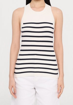 Kobieta ubrana w białą dzianinową bluzkę bez rękawów z czarnymi poziomymi paskami oraz białe spodnie, stojąca ze swobodnie opuszczonymi ramionami.