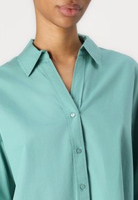 Camicia button-up color teal con colletto e una leggera lucentezza. Presenta una patta con bottoni coordinati, una texture morbida e una vestibilità rilassata.