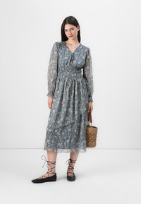 Blommig midiklänning i blått med långa genomskinliga ärmar, smockad midja och V-ringning. Svarta snörskor och en vävd handväska fulländar outfiten.