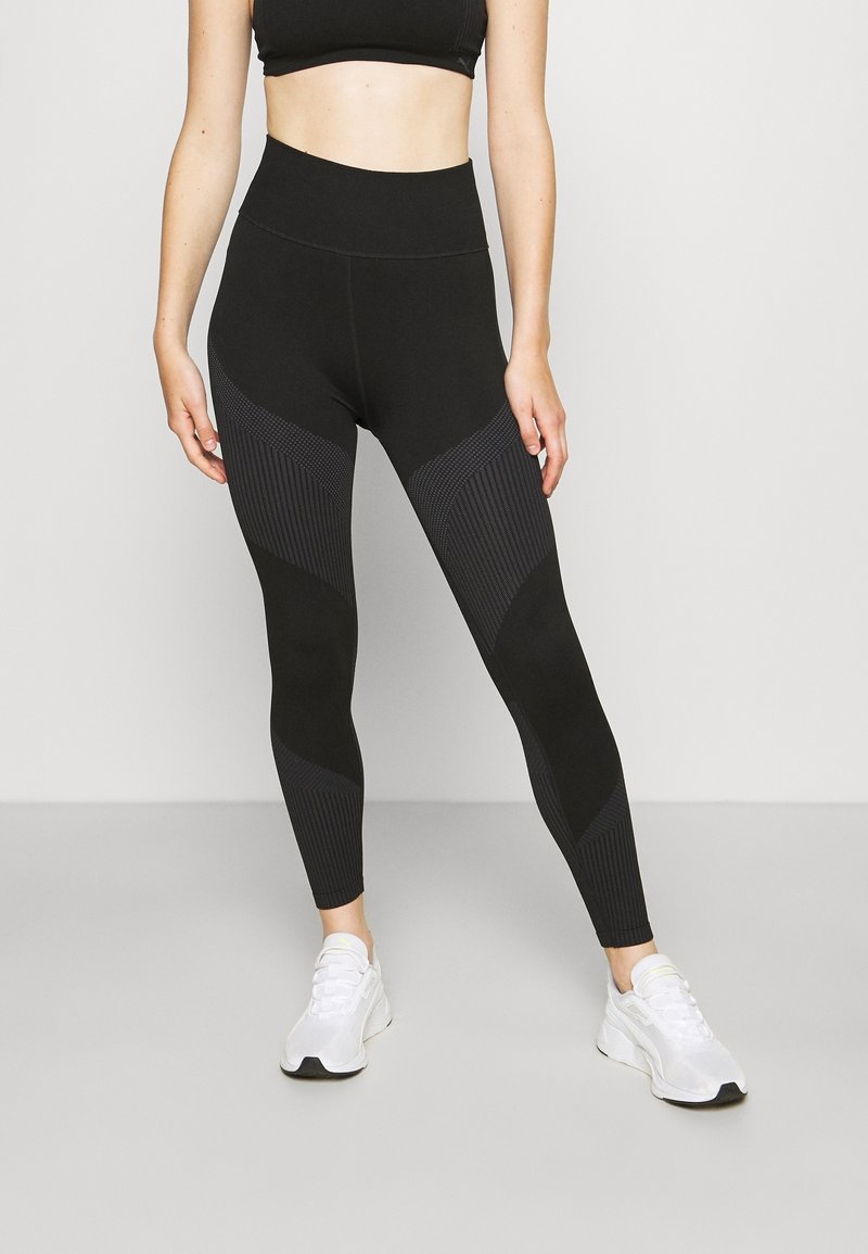 Zwarte high-waisted leggings met geribbelde textuur en mesh accenten. Ze hebben een gevormd ontwerp en zijn gecombineerd met witte sportschoenen.
