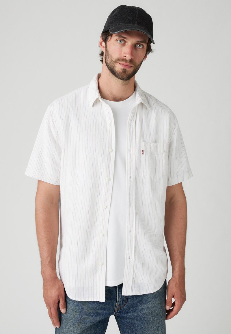 Camicetta a maniche corte di colore bianco con sottili righe verticali, con bottoni frontali e taschino sul lato sinistro, indossata sopra una t-shirt bianca e jeans blu.