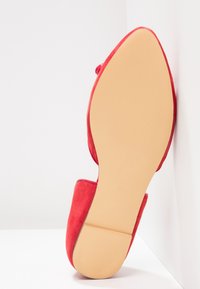 Chaussure plate rouge à bout pointu vue du côté de la semelle beige, positionnée debout contre un mur blanc d'angle.