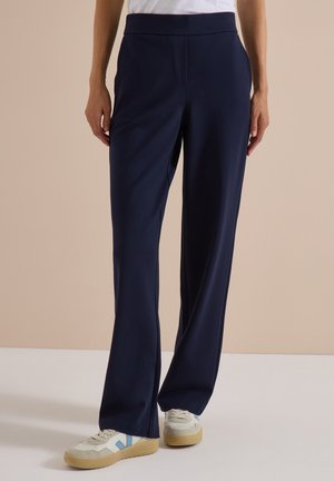 Personne portant un pantalon ample bleu marine foncé et des baskets blanches avec des semelles beige, debout devant un fond beige uni.