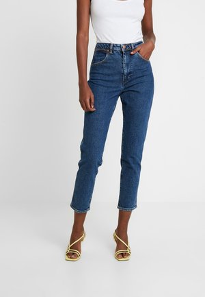 Kobieta nosząca niebieskie dżinsy z wysokim stanem, biały top bez rękawów i żółte sandały na obcasie z paskami, stojąca na jednolitym tle.