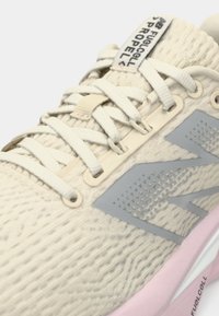 Nærbilde av en beige teksturert løpesko med lys rosa såle, grå "N"-logo og kremfargede lisser knyttet på strikket overdel med "FUELCELL PROPEL"-merke.