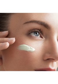 Une crème vert clair est appliquée sur la joue d'une femme. La peau semble lisse et bien entretenue, mettant en valeur la texture et l'hydratation.