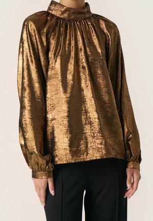 Kvinde iført en langærmet, skinnende bronzefarvet bluse med høj halsudskæring og rynkede detaljer, kombineret med sorte bukser.