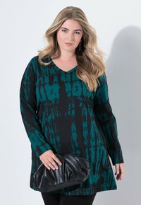 Groene en zwarte lange mouw tuniek met een v-hals, voorzien van een tie-dye patroon. Model houdt een zwarte gestructureerde clutch vast.
