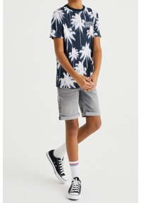 Námořnické tričko s tie-dye designem a bílým vzorem palmy, krátké šedé denimové šortky s ohrnutými nohavicemi, bílé ponožky s červenými a modrými pruhy, černé tenisky.