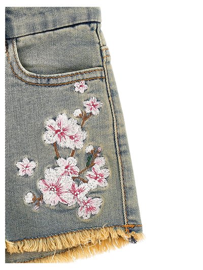 Pantalones cortos de mezclilla con dobladillo deshilachado, que presentan flores de cerezo bordadas en rosa y blanco en la zona del bolsillo delantero.