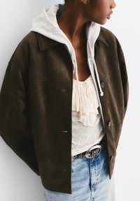 Veste en daim marron avec un sweatshirt gris en dessous, dotée de boutons, d'une coupe décontractée et d'un haut blanc plissé ; associée à un jean en denim clair.