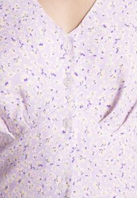 Robe florale lavande avec des motifs violet pâle, crème et verts, avec un col en V et quatre boutons ronds translucides sur le devant.