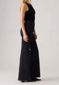 Femme portant un jean noir évasé taille haute et un top noir sans manches, debout de profil devant un fond uni.